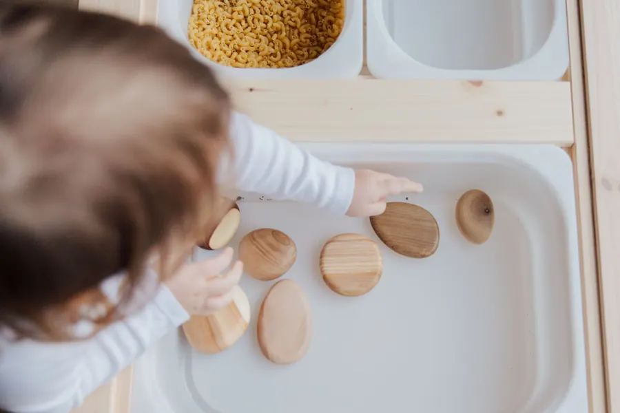 A toddler, seen from behind, sorting smooth wooden pieces in a sensory tray