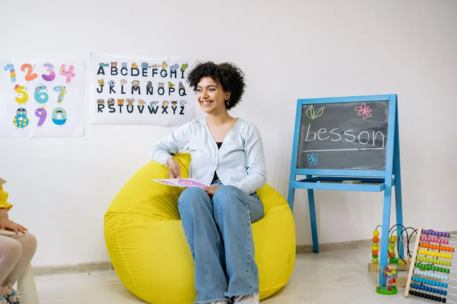 A smiling teacher sitting on a yellow beanbag next to an alphabet poster and a chalkboard easel